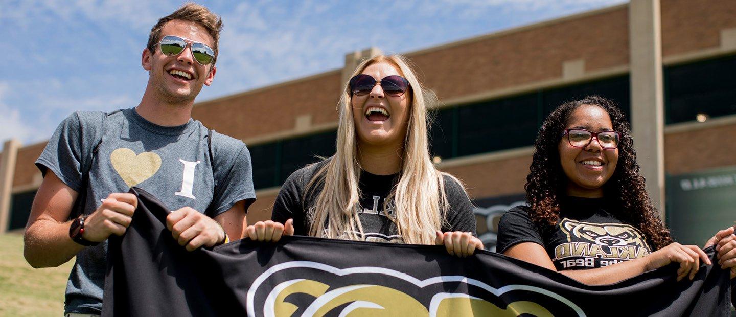 Three O U students holding a black flag with the gold Grizz head mascot on it.